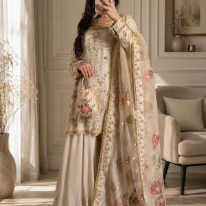 Pode incluir: Um conjunto de túnica de manga comprida e calças largas em tom creme, com bordados dourados intrincados e padrões florais. O traje inclui uma dupatta transparente a condizer com detalhes florais de lantejoulas e uma pequena bolsa redonda.