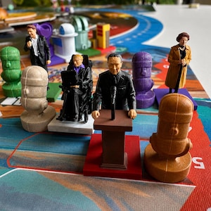 May include: A collection of miniature plastic figurines placed on a colorful board game surface. The set includes figures of people in suits, a person in a wheelchair, and several stylized, solid-colored microphone-shaped game tokens.