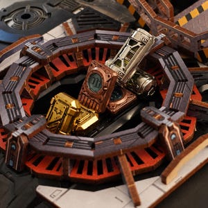 May include: A miniature sci-fi terrain set featuring a circular dark gray and orange platform with metallic copper and gold rectangular containers and a silver cylindrical canister placed in the center, surrounded by industrial structural elements.