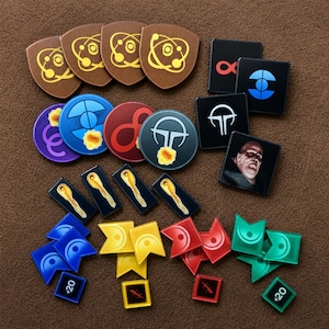 Può includere: Una collezione di gettoni da gioco che include pezzi marroni a forma di scudo con simboli atomici gialli, dischi circolari viola, blu, rossi e grigi, tessere quadrate nere con varie icone e un ritratto, e piccoli segnapunti colorati a forma di bandiera.