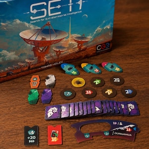 May include: A board game box titled SETI Search for Extraterrestrial Intelligence with a landscape illustration of satellite dishes. Included are various game components: colorful shaped tokens, circular dark markers, and rectangular purple tiles.