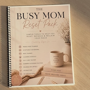 Op de afbeelding: Een spiraalgebonden planner getiteld "The Busy Mom Reset Pack" met een zachtroze omslag. De tekst op de omslag luidt "Eenvoudige tools om u te helpen resetten, herfocussen en uw vrede terug te winnen". Een mok, boeken en een pen zijn ook zichtbaar.