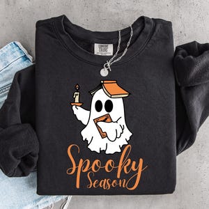 Peut inclure: Un sweat-shirt noir à manches longues avec un motif représentant un fantôme blanc portant un livre sur la tête, tenant une bougie allumée et un autre livre. Le texte "Spooky Season" est imprimé en orange sous le fantôme. Une chaîne en argent.