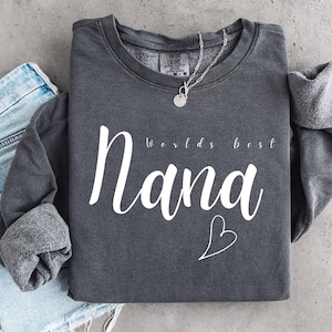 May include: A charcoal grey crewneck sweatshirt featuring the white text "worlds best Nana" in a script font, accompanied by a simple white heart outline. The garment has a fleece-lined interior and a silver-toned necklace resting on the collar.