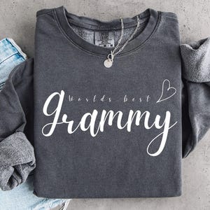 Może przedstawiać: Ciemnoszara bluza z okrągłym dekoltem z białym napisem "worlds best Grammy" wykonanym mieszanką czcionek pisanych i bezszeryfowych, z małym białym serduszkiem. Odzież posiada polarowe wnętrze oraz widoczną metkę producenta.