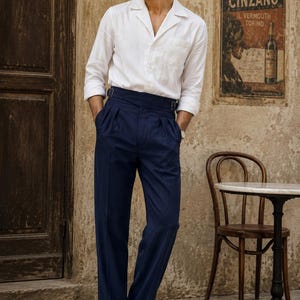 Può includere: Una camicia bianca a maniche lunghe con colletto a revers e taschino sul petto, abbinata a pantaloni blu navy a vita alta con doppia pince, cinturini laterali regolabili e risvolto, indossata con mocassini in pelle marrone scuro.