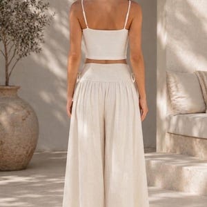 Pode incluir: Um conjunto de duas pe&ccedil;as composto por uma camisola curta de cor creme com al&ccedil;as finas e cal&ccedil;as a condizer de cintura subida e perna larga. As cal&ccedil;as apresentam uma cintura franzida com detalhes de la&ccedil;os laterais e um corte largo.
