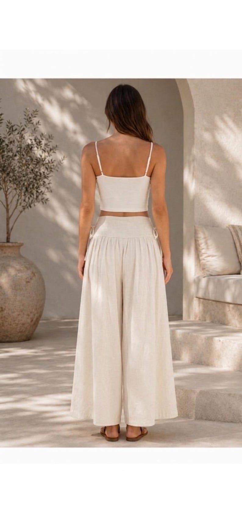 Pode incluir: Um conjunto de duas pe&ccedil;as composto por uma camisola curta de cor creme com al&ccedil;as finas e cal&ccedil;as a condizer de cintura subida e perna larga. As cal&ccedil;as apresentam uma cintura franzida com detalhes de la&ccedil;os laterais e um corte largo.