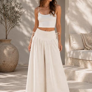 Pode incluir: Um top curto sem mangas branco combinado com cal&ccedil;as creme de cintura subida e perna larga. As cal&ccedil;as apresentam uma cintura franzida com la&ccedil;os laterais ajust&aacute;veis e uma silhueta fluida e larga que se estende at&eacute; aos tornozelos.