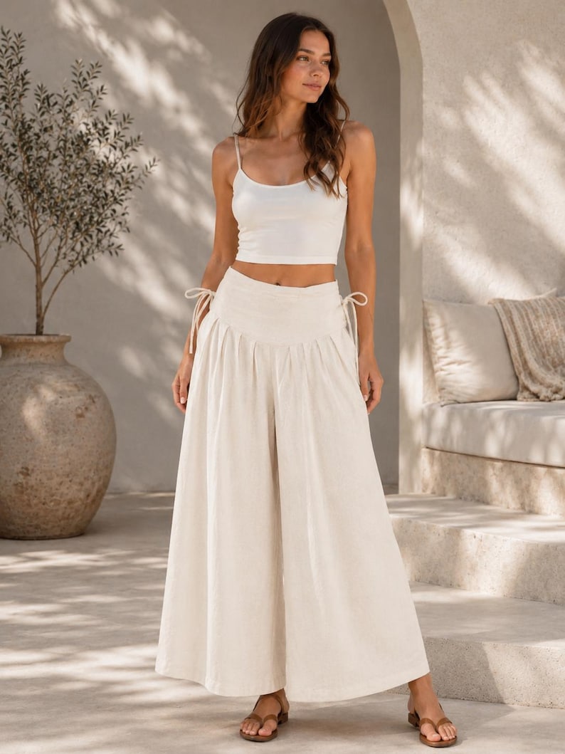 Pode incluir: Um top curto sem mangas branco combinado com cal&ccedil;as creme de cintura subida e perna larga. As cal&ccedil;as apresentam uma cintura franzida com la&ccedil;os laterais ajust&aacute;veis e uma silhueta fluida e larga que se estende at&eacute; aos tornozelos.