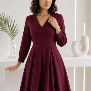May include: A deep burgundy midi dress featuring a V-neckline, long sleeves with buttoned cuffs, and a front button placket. The bodice is fitted with a defined waistline, transitioning into a pleated, flared skirt that falls below the knees.