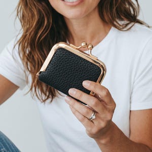 May include: A small black coin purse with a textured pebbled leather finish and a gold-toned metal frame. The purse features a classic kiss-lock closure with two round gold-toned beads at the top, providing a secure and functional design.
