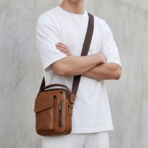 May include: A tan crossbody bag featuring a top handle, a front flap pocket, and a vertical zipped compartment. The bag includes a dark brown adjustable shoulder strap with silver-toned metal hardware and a small circular grommet detail.