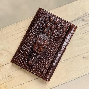 May include: A dark brown bifold wallet featuring a prominent embossed crocodile skin texture. The front surface displays a detailed, raised pattern resembling the head and scales of a crocodile, with a glossy finish and a folded edge design.