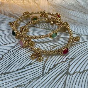 May include: Two gold-toned twisted metal bangles featuring small, round, multi-coloured cabochon stones in shades of pink, green, and red. The bangles are accented with small, dangling gold-toned bell-shaped charms attached at intervals along the band.