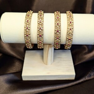 May include: Four gold-toned metal bangles featuring an intricate filigree floral pattern. Each bangle is embellished with small, round, deep pink stones set in the centre of each flower, creating a repeating decorative design across the surface.