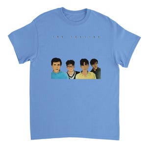May include: A light blue short-sleeve t-shirt featuring a graphic print of four individuals and the text "the feelies" centered above them. The individuals are depicted in a realistic art style with varied clothing colors including blue, yellow, and dark tones.