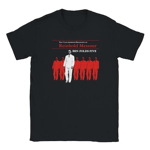 Op de afbeelding: Een zwart t-shirt met korte mouwen en een grafische print met de tekst The Unauthorized Biography of Reinhold Messner en Ben Folds Five. Het ontwerp toont een centrale figuur in een wit pak, omringd door zes identieke figuren in rode pakken.