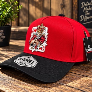 May include: A red and black baseball cap featuring a graphic of a King of Hearts playing card on the front. The black brim displays a silver sticker with the text KAMEL PREMIUM QUALITY HEADWEAR 804. A black hang tag with the KAMEL logo is attached.