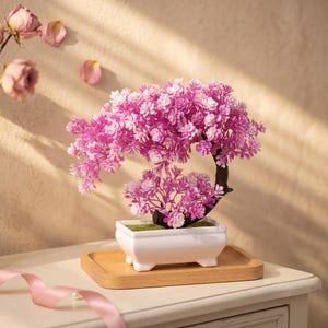May include: A small artificial bonsai tree with vibrant pink and white flowers, featuring a dark brown curved trunk. The tree is planted in a rectangular white ceramic pot with four small feet, resting on a light-colored wooden tray.