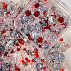 May include: A collection of assorted beads in shades of red, clear, and iridescent silver. The mix includes faceted crystal-style beads, smooth round spheres, and small seed beads, with a portion contained in a clear square plastic container.
