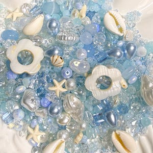 May include: A collection of assorted beads in various shades of blue, clear, and white. The mix includes small round beads, faceted crystals, pearlescent spheres, white flower-shaped charms, and natural-looking seashell and starfish accents.