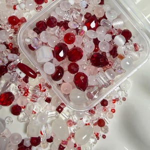 May include: A collection of assorted beads in shades of red, white, and clear, contained within a square transparent plastic storage box. The beads vary in shape, including round, faceted, cube, and teardrop styles, with some featuring iridescent finishes.