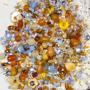 May include: A collection of assorted beads in shades of amber, gold, yellow, and light blue. The mix includes various shapes such as round, faceted, teardrop, and flower-shaped beads, featuring translucent, opaque, and metallic finishes.
