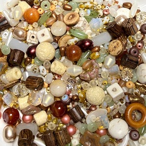 May include: A large collection of assorted beads in various shapes, sizes, and textures. The mix includes wooden cylinders, smooth round stones, faceted glass, iridescent pearls, and matte ceramic cubes in shades of brown, cream, and green.