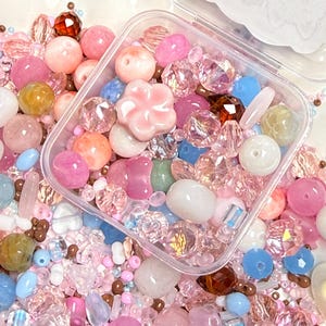Könnte beinhalten: Eine Sammlung verschiedener Perlen in einem durchsichtigen Kunststoffbehälter und verstreut darum herum. Die Perlen variieren in Form, Größe und Farbe, darunter Rosa-, Blau-, Weiß- und Brauntöne, mit facettierten, glatten und blumenförmigen Stücken.