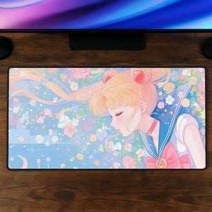 Op de afbeelding: Een rechthoekige muismat met een anime-stijl illustratie van een personage met blond haar, gekleed in een matrozenuniform. De achtergrond is lichtblauw met bloemen- en steraccenten. De mat ligt op een houten oppervlak.