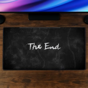Peut inclure: Un tapis de bureau rectangulaire noir présentant un fond sombre et texturé avec de légères illustrations fantomatiques de personnages et de structures. Les mots The End sont imprimés en blanc, dans une police manuscrite et usée.