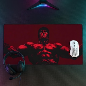 Puede incluir: Una alfombrilla de escritorio rectangular con una ilustración en rojo oscuro y negro de un personaje musculoso de pelo corto. La alfombrilla se sitúa bajo un ratón de ordenador blanco con patrón de panal y unos auriculares negros.