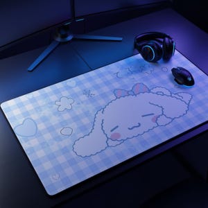 Puede incluir: Una alfombrilla de escritorio rectangular con un patrón de cuadros azul claro y blanco y una gran ilustración de un personaje blanco y esponjoso. El diseño incluye mejillas rosas, un lazo rosa y contornos de corazones y flores, con texto japonés.