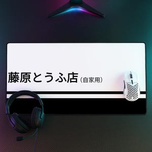 May include: A rectangular desk mat featuring a white top section with black Japanese text reading 藤原とうふ店 (自家用) and a black bottom section separated by a thin white line. A white honeycomb-patterned gaming mouse and black wired headphones are placed on the mat.