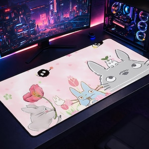 Peut inclure: Un tapis de souris rose avec des illustrations de personnages du film d'animation "Mon Voisin Totoro". Le design comprend Totoro, des lapins bleus et d'autres esprits de la forêt, avec des accents floraux et de feuilles. Le tapis de souris est rectangulaire.