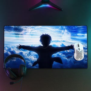 Puede incluir: Una alfombrilla de escritorio rectangular con un gráfico impreso de la silueta oscura de una persona con los brazos extendidos contra un cielo azul brillante lleno de nubes. Un ratón de ordenador blanco con patrón de panal y auriculares negros con cable están colocados encima.