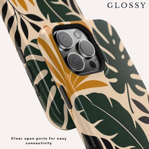 May include: A glossy phone case featuring a botanical pattern of dark green, mustard yellow, and cream leaves. The case has a triple camera cutout, black side buttons, and the text GLOSSY and Clear open ports for easy connectivity.