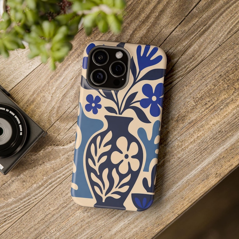 May include: A smartphone case featuring a cream background with a dark blue vase containing a floral arrangement. The design includes stylized blue flowers, leaves, and abstract shapes in various shades of blue, covering the entire back surface.