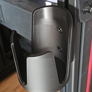 May include: A black, curved metal bracket with a cylindrical holder. The bracket is attached to a larger black structure with two screws. The holder has a smooth, matte finish and is designed to hold an object.
