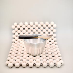 May include: A silver-coloured metal bowl sits on a square mat constructed from rows of small, round, off-white wooden beads. A two-toned spoon with a dark handle and a light-coloured bowl rests horizontally across the top of the metal bowl.