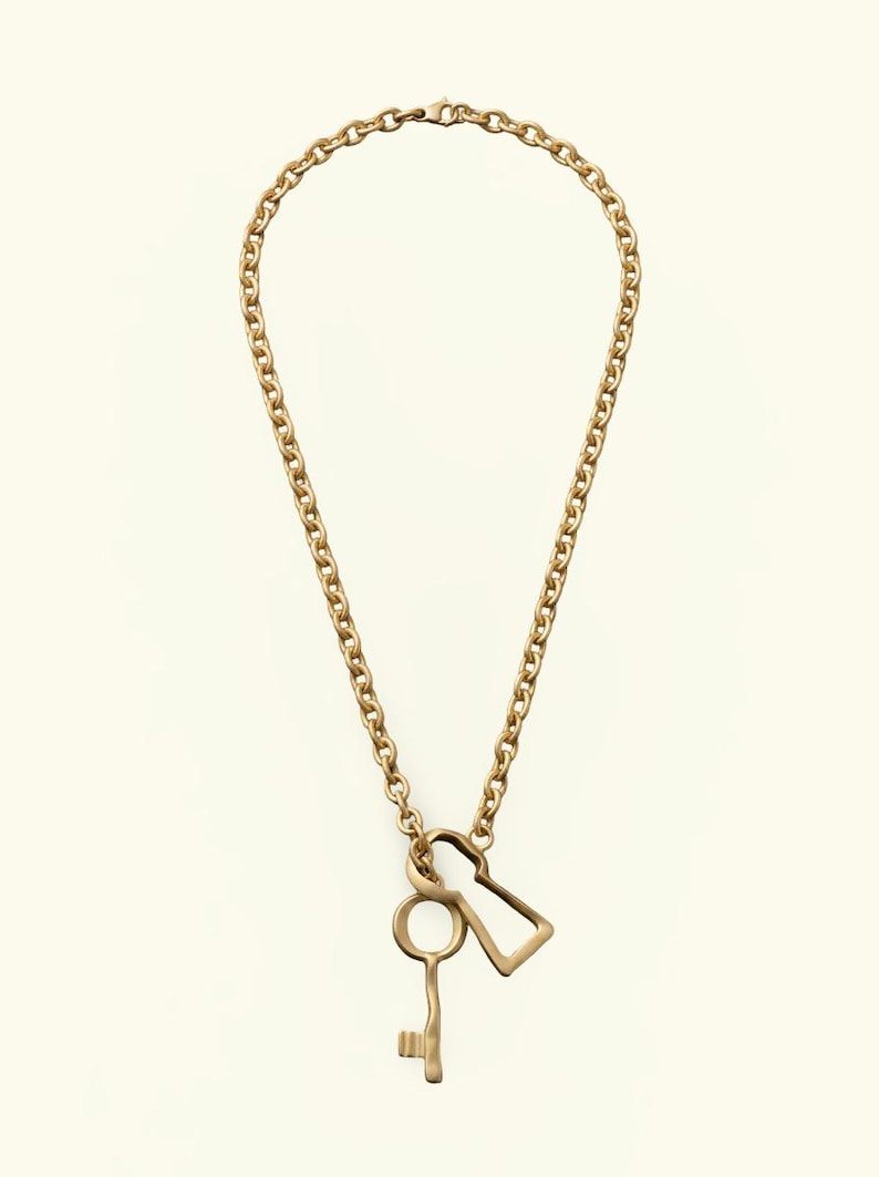 May include: A gold-toned necklace featuring a thick chain link design. Suspended from the chain are two distinct metal pendants: one shaped like a traditional skeleton key and the other shaped like a classic keyhole silhouette, both in gold.