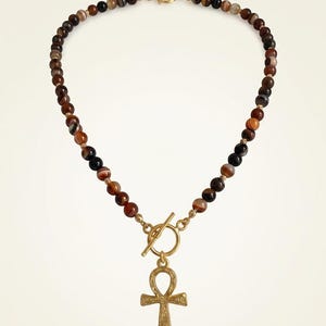May include: A beaded necklace featuring round, multi-colored brown and black banded agate beads interspersed with small gold-toned spacers. The necklace includes a gold-toned toggle clasp and a central gold-toned ankh pendant with textured detail.