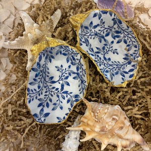 May include: Two oyster shells featuring a white interior with a blue botanical leaf pattern and gold-painted edges, accompanied by a starfish, a spiral seashell, and a small purple shell, all arranged on a bed of shredded brown paper.