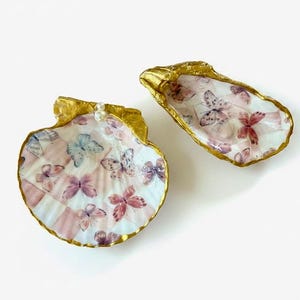 May include: Two decorative seashell dishes featuring a delicate butterfly pattern in shades of pink, blue, and purple. The edges of the shells are painted with a metallic gold finish, and the round shell includes a small white pearl accent.