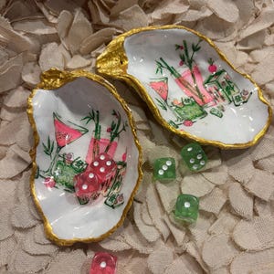 Può includere: Due conchiglie di ostrica con bordi dipinti in oro, caratterizzate da illustrazioni dipinte a mano di bambù e tessere del mahjong in rosa e verde. Le conchiglie sono accompagnate da quattro dadi traslucidi a sei facce, due rosa e due verdi, con puntini bianchi.