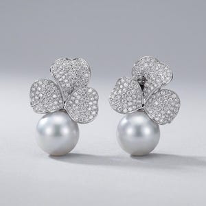 May include: A pair of stud earrings featuring a large, round, lustrous white pearl base topped with a three-petaled flower design. Each petal is densely encrusted with small, sparkling clear stones set in a bright, polished silver-toned metal.