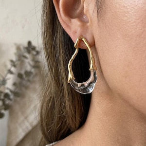May include: A drop earring featuring an irregular, organic-shaped gold metal frame that transitions into a clear, smooth, translucent resin base. The design has a textured, molten appearance on the gold portion and a curved, teardrop-like silhouette.
