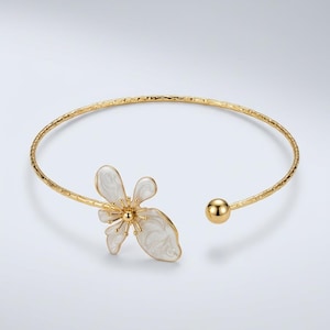 May include: A gold-tone open cuff bracelet featuring a textured band and a white enamel flower accent on one end. The opposite end of the bracelet terminates in a smooth, polished gold-tone sphere. The flower has delicate gold-tone stamens.