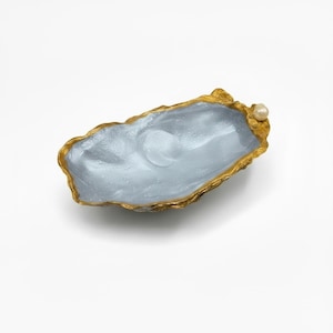 May include: An oyster shell dish with a shimmering light blue interior and a textured gold-painted rim. A single small white pearl is attached to one end of the shell, accentuating the organic, irregular shape of the decorative bowl.